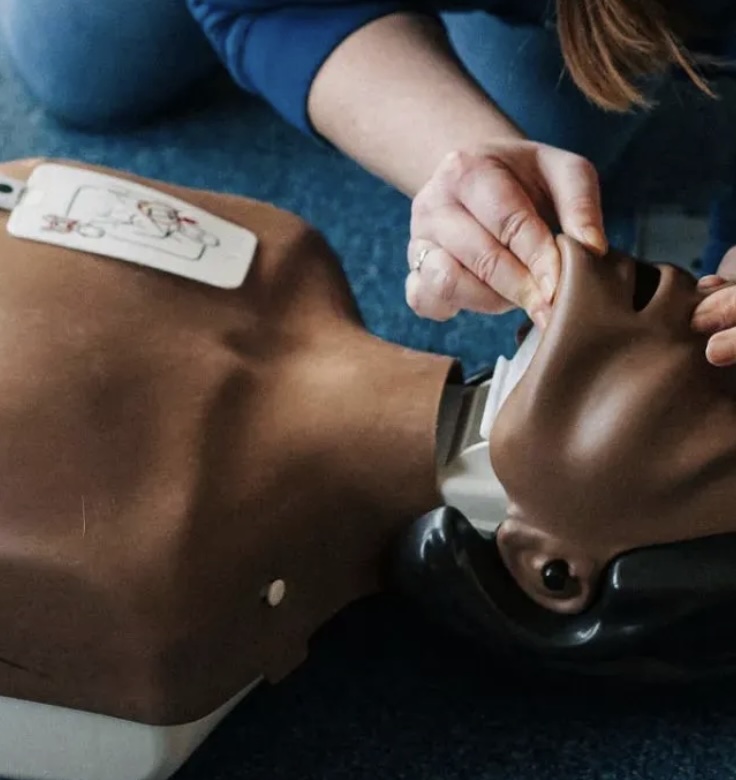 A person practicing first aid on a CPR training manikin, demonstrating opening the airway by placing the index and middle fingers of one hand on the manikin's chin.