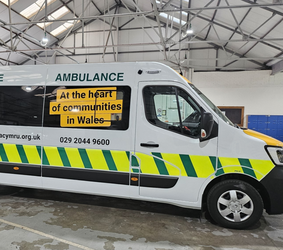 The side of ambulance parked in a garage. It is white, yellow and green and has the strapline 'At the heart of communities in Wales' on the side.