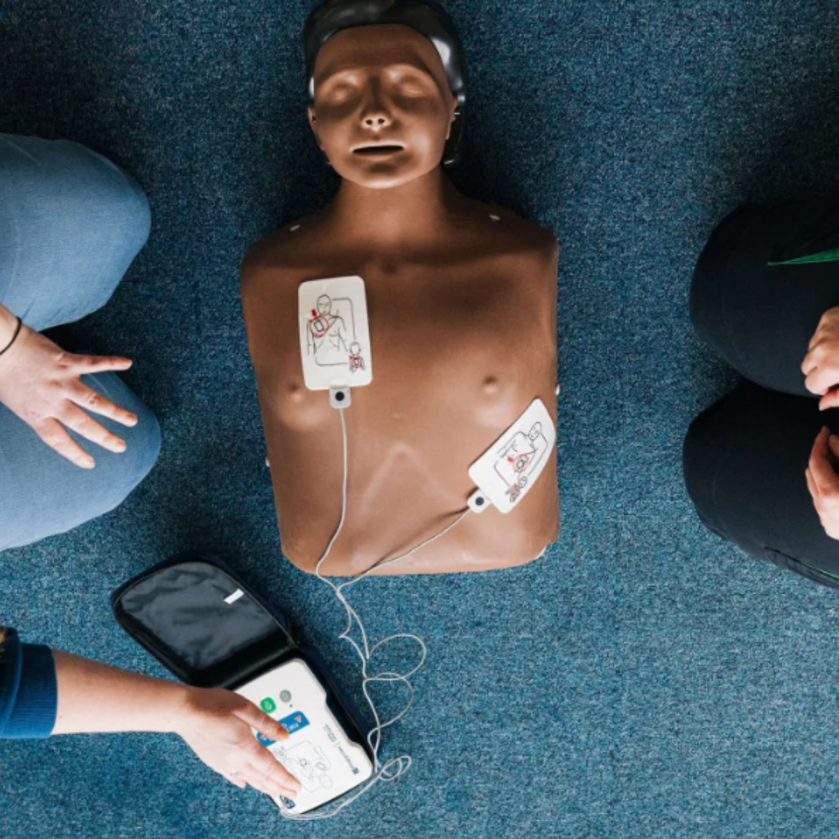 A CPR training manikin laid on the carpet floor. Someone is knelt down next to the manikin and is practicing placing the pads on to the 'casualty’s' chest by applying them in the positions shown on the device.