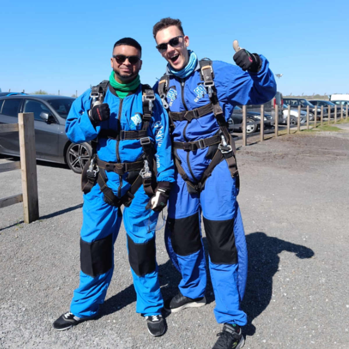 Az and Nkosi posing outside before their skydive, standing next to each other in blue skydive overalls. Both are wearing sunglasses and harnesses over their overalls.
