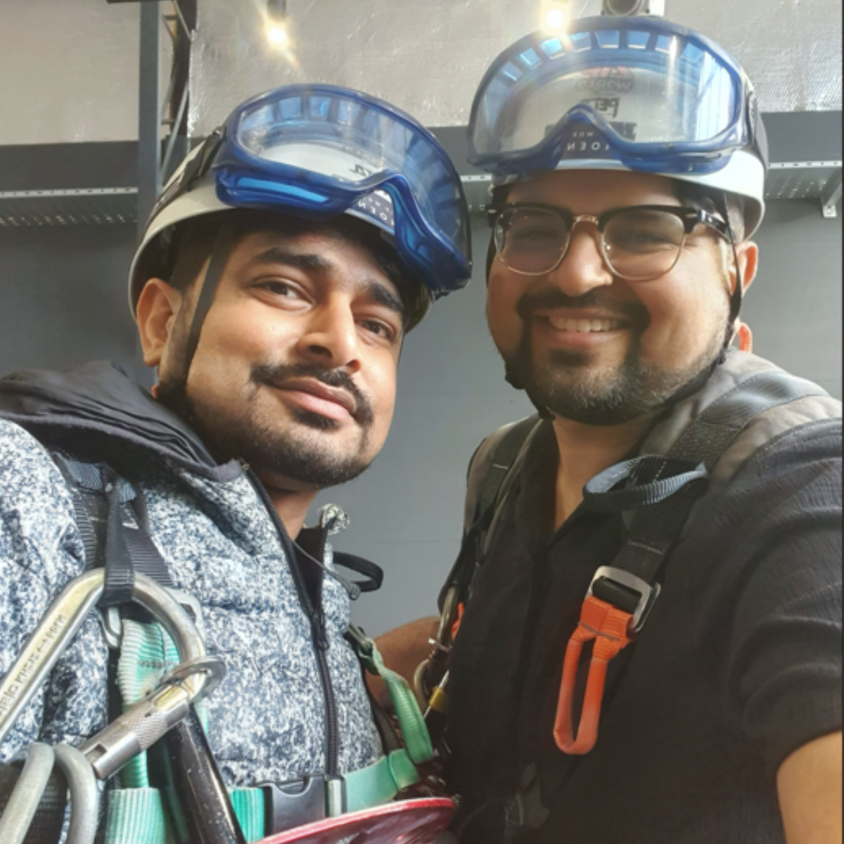Dev and Priyank smiling while taking a selfie before their zipline challenge. They are both wearing hard hats, goggles, and safety harnesses.