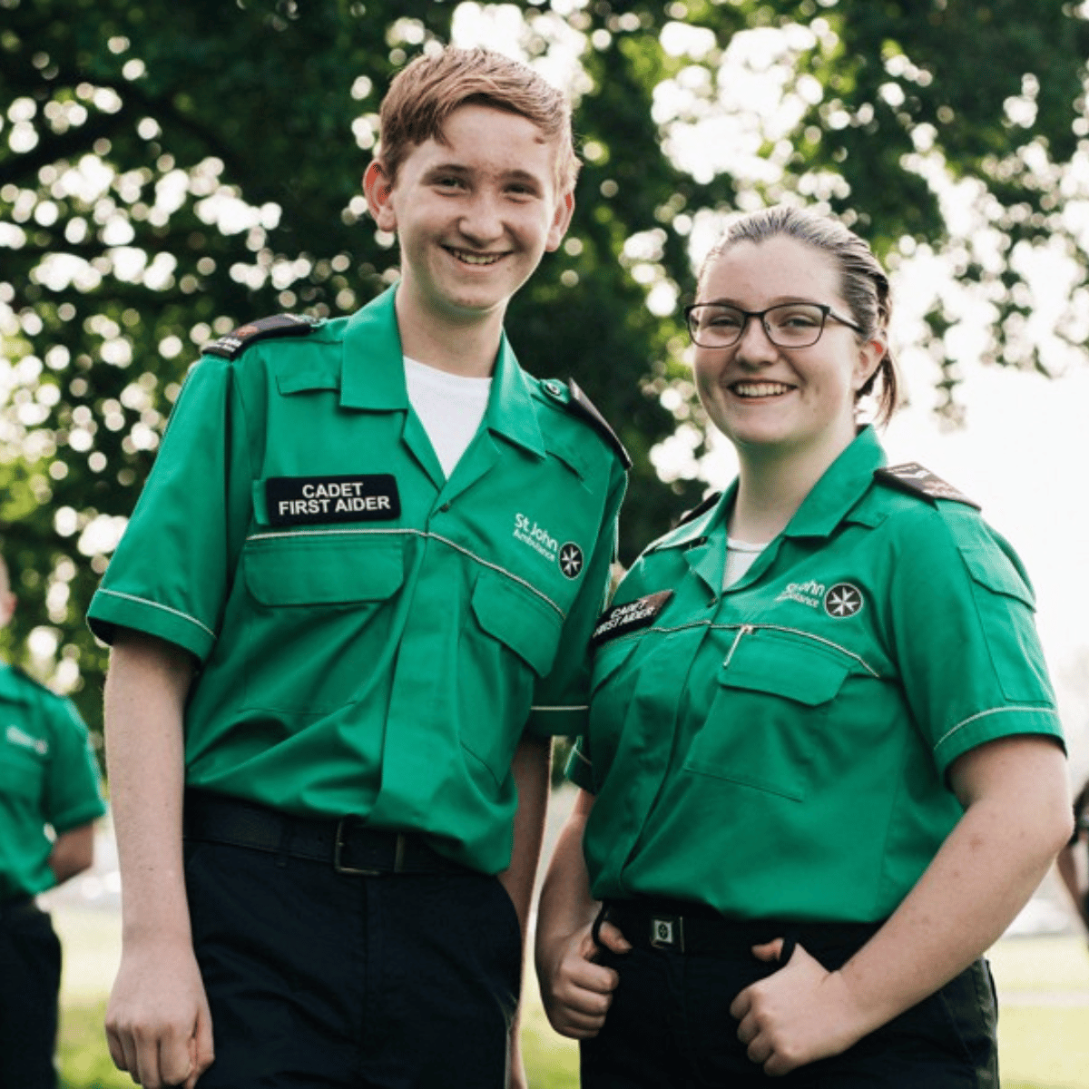 Two cadet first aiders standing outdoors, smiling, wearing green St John Ambulance Cymru uniforms with the logo and a ‘Cadet First Aider’ badge on their shirts