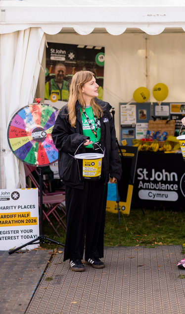 St John Ambulance Cymru staff member wearing a green St John Ambulance Cymru t-shirt and coat, holding a donations bucket. She is standing in front of an event tent and behind her is a table with St John Ambulance Cymru merch on and a table cloth with our logo.
