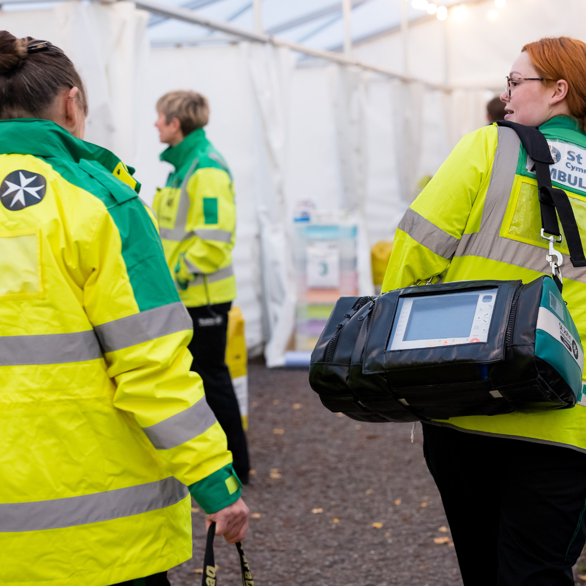 St John Ambulance Cymru volunteers are walking through the medical tent at the Cardiff Half Marathon, carrying medical equipment and talking to each other. There are three volunteers, all wearing bight green high visibility coats.