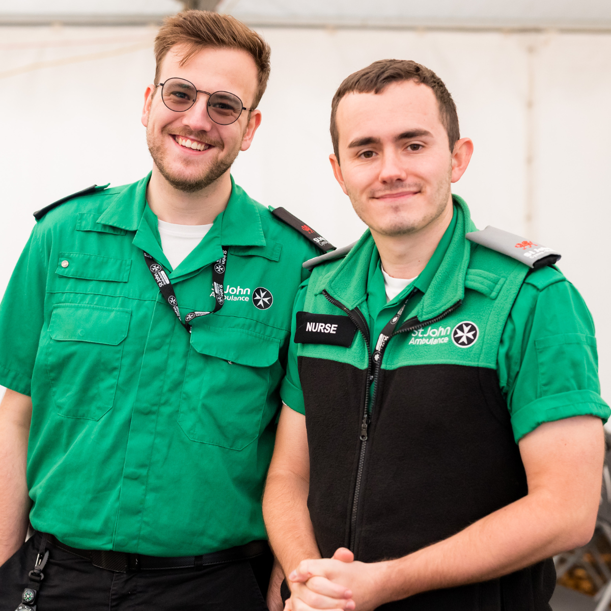 Two St John Ambulance Cymru volunteers smiling next to each other. Both wearing our green uniform. One of them, is wearing a badge with the wording 'Nurse'. They are standing in the medical tent at the Cardiff Half Marathon with the white tent wall in the background.