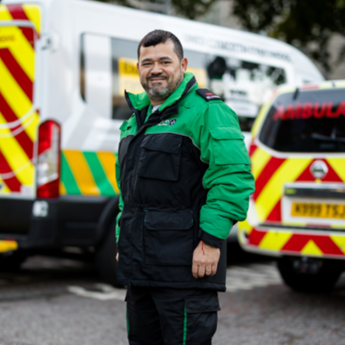 St John ambulance Cymru volunteer wearing green uniform. He is standing in car park with Ambulances behind him. He is smiling and has a beard.