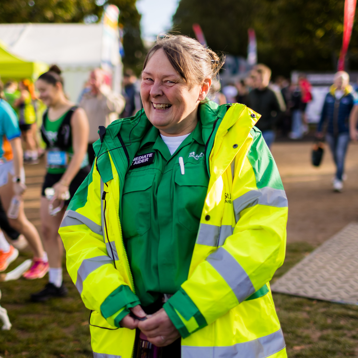 St John Ambulance Cymru volunteer smiling at the Cardiff Half Marathon. She is wearing a bright yellow high visibility coat and has a badge showing she is an immediate first aider.