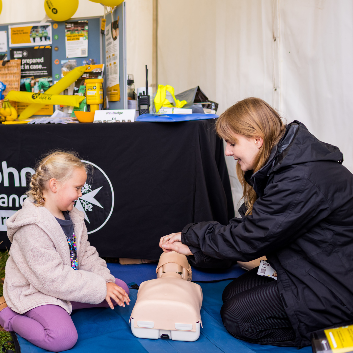 St John Ambulance Cymru staff teaching a little girl how to do CPR on a manikin. They are in a booth at an event. The staff member is wearing a black coat and is demonstrating how to give chest compressions. In the background, is a table which has informational materials on and a table cloth with the St John Ambulance Cymru logo.