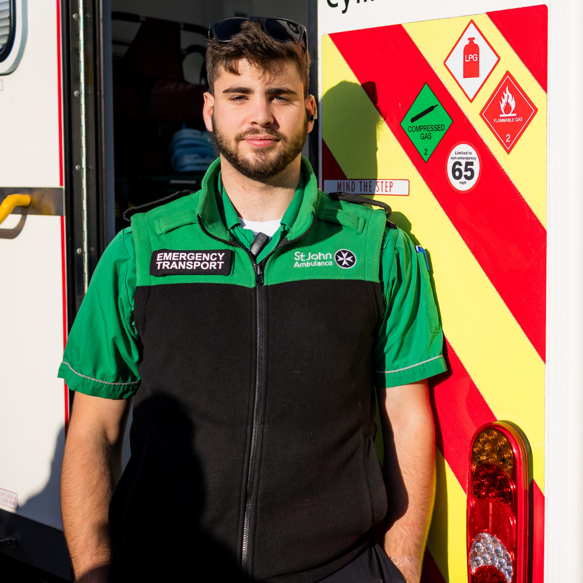 St John Ambulance Cymru staff member standing outside by Ambulance backdoor, wearing green St John Ambulance uniform and smiling. He has a badge on his uniform which reads Emergency Transport, as well as the logo.