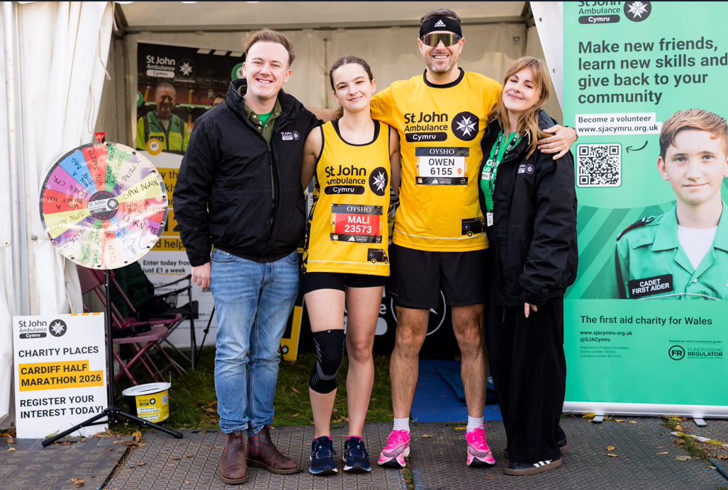 Two St John Ambulance Cymru staff members standing with two runners from the Cardiff Half Marathon. The runners are wearing the St John Ambulance Cymru vest, which fundraisers for St John Ambulance Cymru wear for the race. They are all smiling.