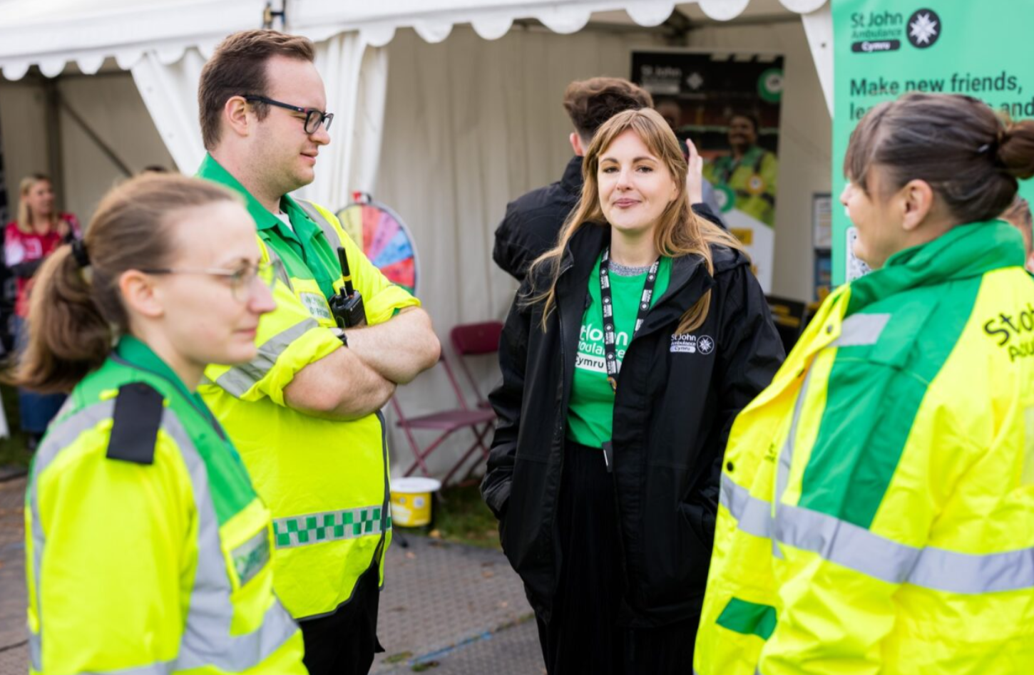A group of four people, three wearing bright yellow and green high-visibility jackets with the St John Ambulance Cymru logo on them, stand and chat outdoors near a white tent at an event. One person in the centre, wearing a black jacket over a green shirt with a lanyard, is looking directly at the camera with a slight smile.