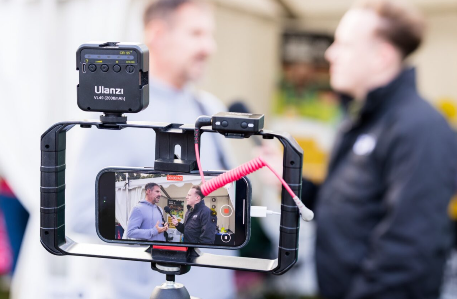 Traitors finalist and St John Ambulance Cymru ambassador Andrew Jenkins is being interviewed by a St John Ambulance Cymru staff member. They are standing in front of the St John Ambulance Cymru tent at the Cardiff Half Marathon. Behind him, in the tent are banners with the St John Ambulance Cymru logo. They are in mid conversation and Andrew is talking into a microphone. They are being filmed by a phone on a tripod.